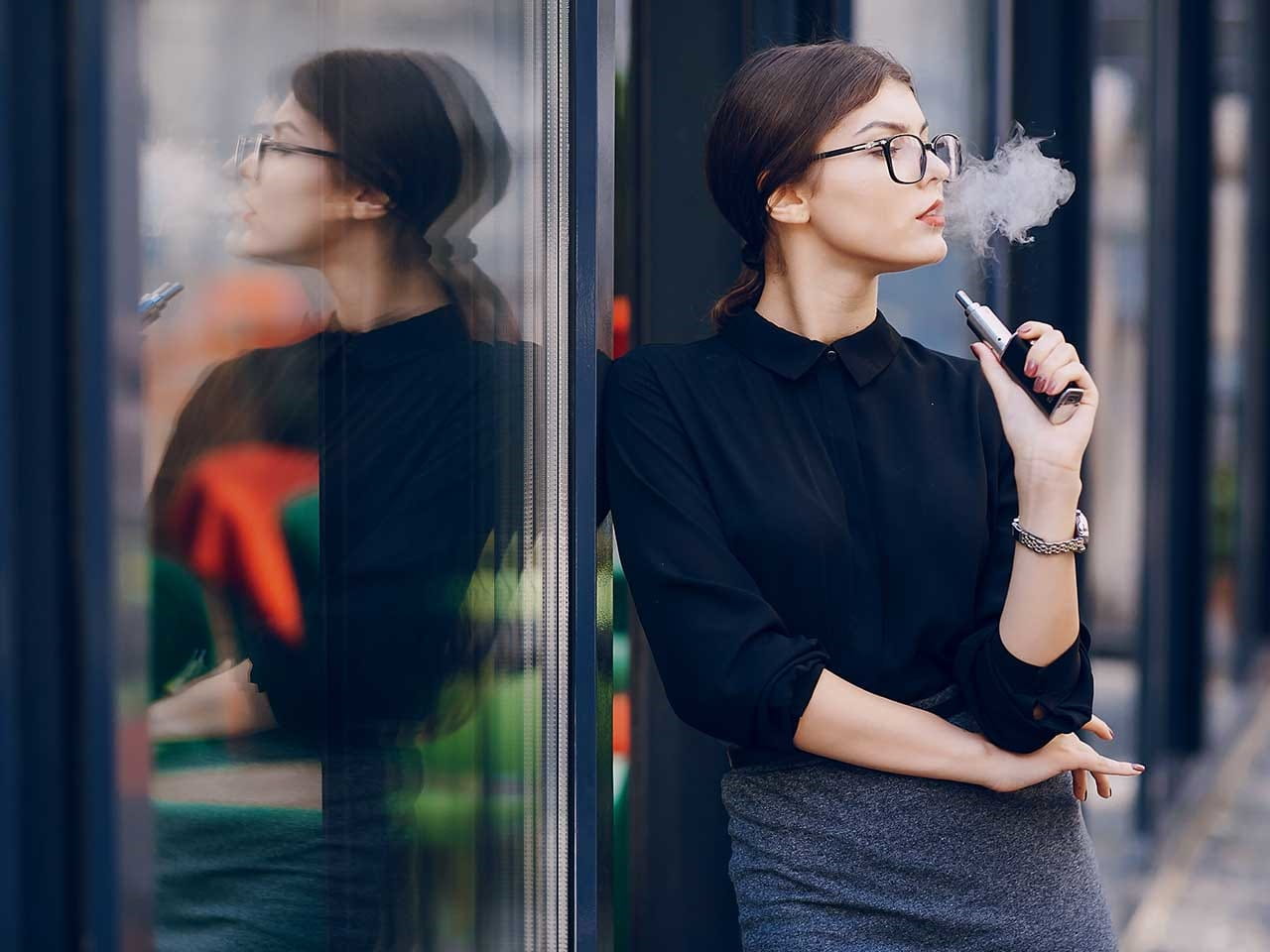 Woman smoking a vape whilst standing against a glass window 